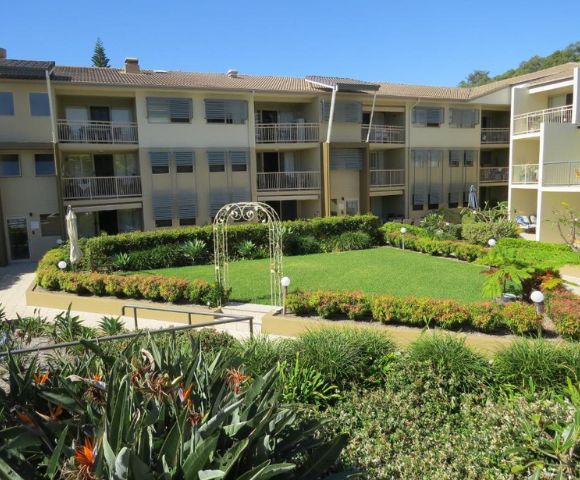 A three-story apartment building surrounds a neatly manicured central garden with lush greenery and a decorative archway, conveying a serene atmosphere.