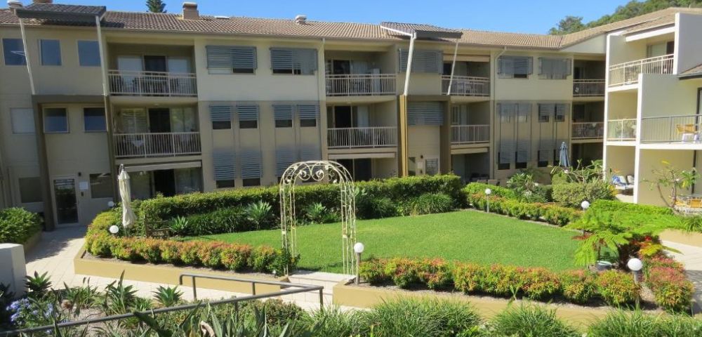 A three-story apartment building surrounds a neatly manicured central garden with lush greenery and a decorative archway, conveying a serene atmosphere.
