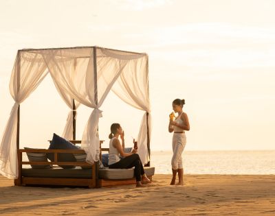 Two people relax on a covered beach bed at sunset, enjoying drinks. One is seated, while the other stands. The sky is golden, creating a serene atmosphere.