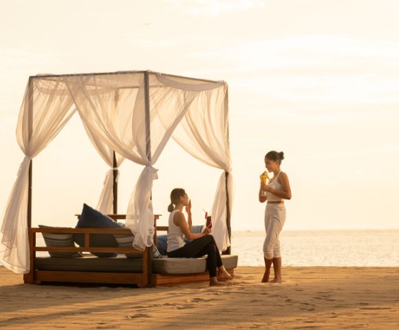 Two people relax on a covered beach bed at sunset, enjoying drinks. One is seated, while the other stands. The sky is golden, creating a serene atmosphere.