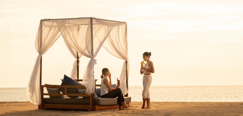 Two people relax on a covered beach bed at sunset, enjoying drinks. One is seated, while the other stands. The sky is golden, creating a serene atmosphere.