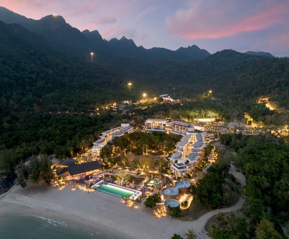 Aerial view of a well-lit resort nestled in lush green hills at dusk. The complex includes a large pool, winding pathways, and surrounding mountains.