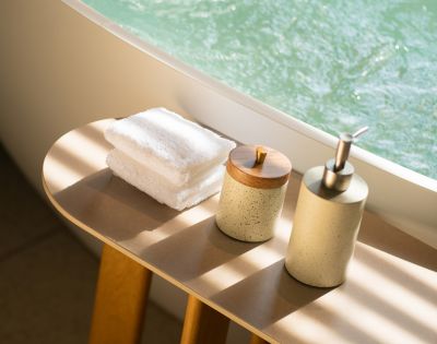 Sunlit bathroom scene with white towels stacked on a wooden table. A soap dispenser and lidded container create a serene, spa-like atmosphere.