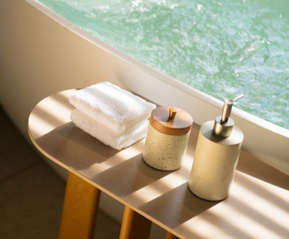 Sunlit bathroom scene with white towels stacked on a wooden table. A soap dispenser and lidded container create a serene, spa-like atmosphere.