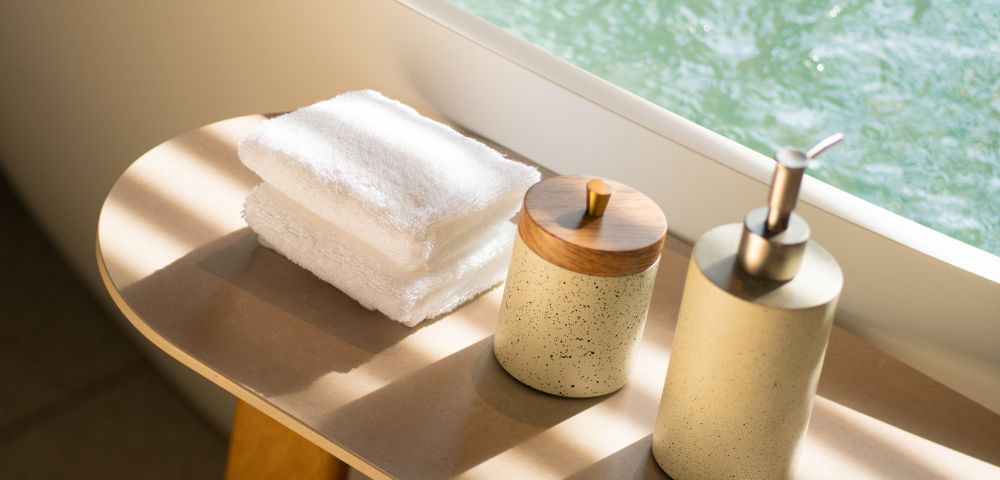 Sunlit bathroom scene with white towels stacked on a wooden table. A soap dispenser and lidded container create a serene, spa-like atmosphere.