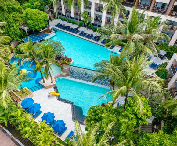 Aerial view of a tropical resort pool surrounded by lush palm trees and greenery. Blue umbrellas and sun loungers line the pool, creating a relaxed atmosphere.