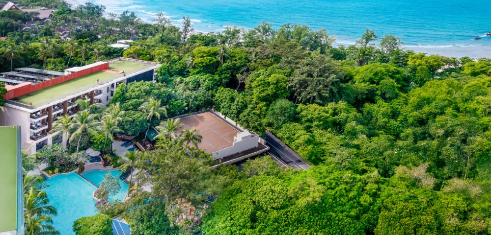 Aerial view of a resort surrounded by lush green trees near a turquoise beach. Features a large pool, palm trees, and a coastal landscape. Serene ambiance.