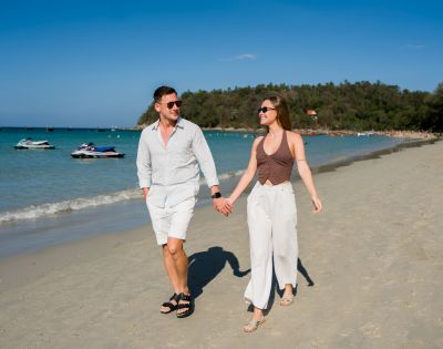A couple holding hands and smiling while walking on a sunny beach, with clear blue water, jet skis, and a forested hill in the background.