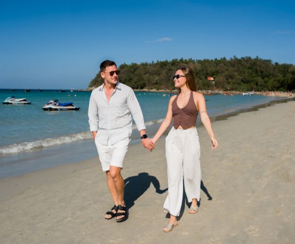 A couple holding hands and smiling while walking on a sunny beach, with clear blue water, jet skis, and a forested hill in the background.