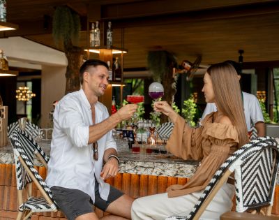 A man and woman sit at a stylish bar, smiling and clinking colorful cocktails. The setting is modern and relaxed, with bottles on display behind them.