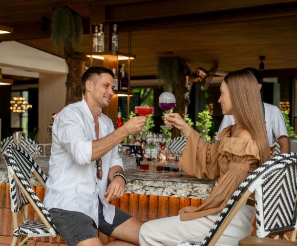 A man and woman sit at a stylish bar, smiling and clinking colorful cocktails. The setting is modern and relaxed, with bottles on display behind them.
