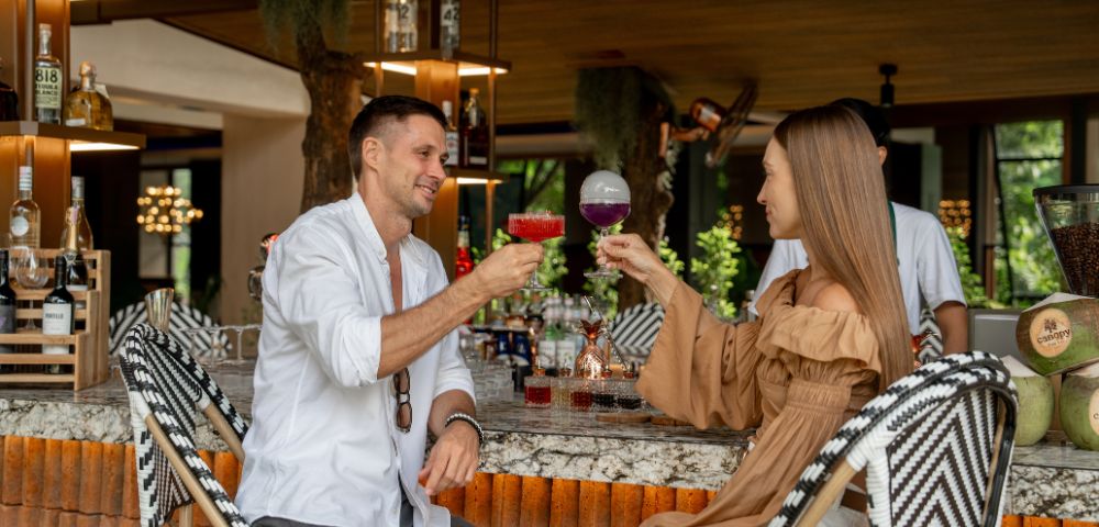 A man and woman sit at a stylish bar, smiling and clinking colorful cocktails. The setting is modern and relaxed, with bottles on display behind them.