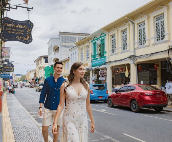 A couple strolls down a vibrant street lined with colorful, historic buildings and signs. The scene conveys a lively, cheerful atmosphere.