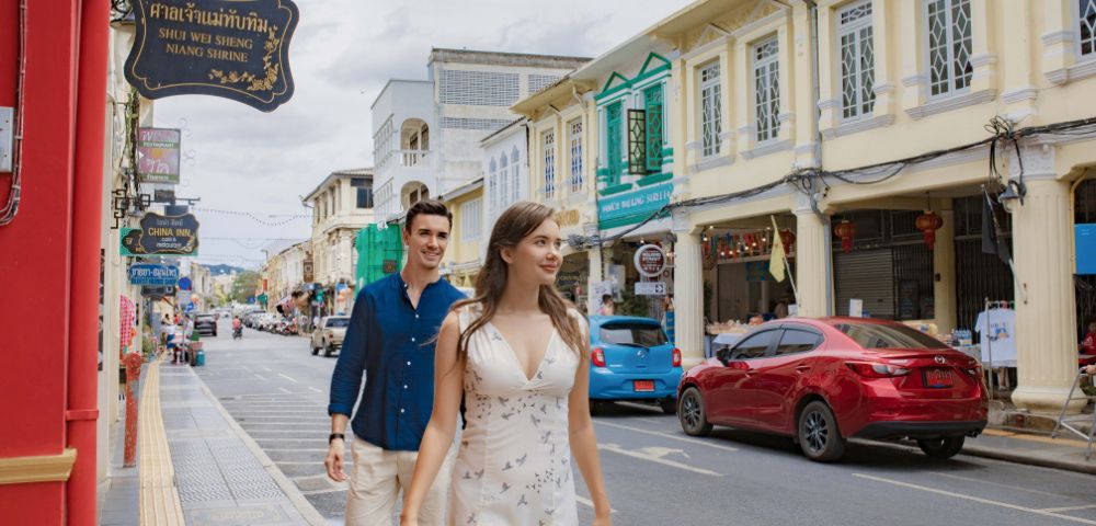 A couple strolls down a vibrant street lined with colorful, historic buildings and signs. The scene conveys a lively, cheerful atmosphere.