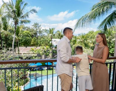 A family of three stands on a balcony, overlooking a lush tropical scene with palm trees and a pool. The mood is relaxed and joyful.