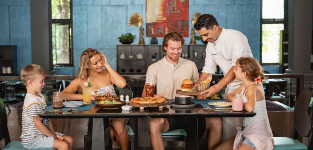 A cheerful family of four sits at a restaurant table filled with food and drinks. A waiter serves a burger, creating a warm and welcoming atmosphere.