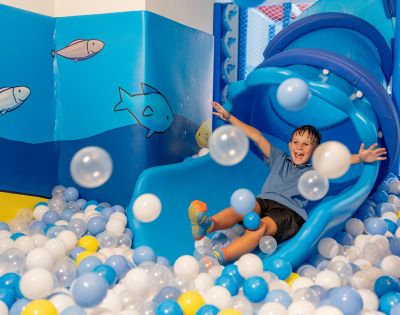 A joyful child slides down a blue slide into a ball pit filled with blue, white, and yellow balls. The room is ocean-themed with fish on the walls.