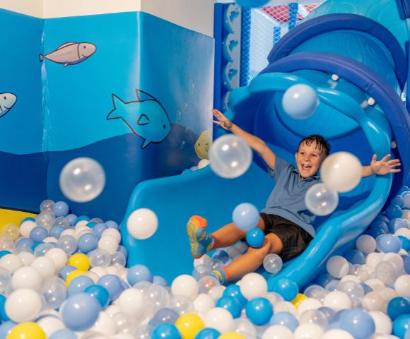 A joyful child slides down a blue slide into a ball pit filled with blue, white, and yellow balls. The room is ocean-themed with fish on the walls.