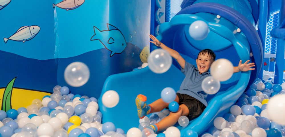 A joyful child slides down a blue slide into a ball pit filled with blue, white, and yellow balls. The room is ocean-themed with fish on the walls.