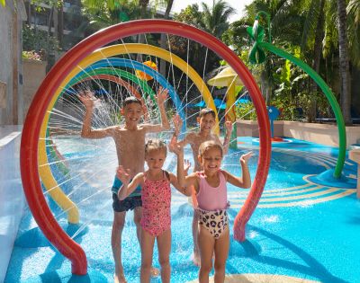 Children joyfully play in a water park, standing under colorful, rainbow-shaped sprinklers. They are smiling, with arms raised amidst splashing water.