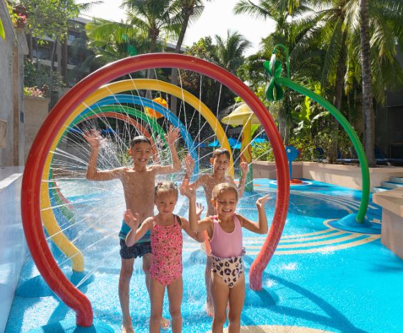Children joyfully play in a water park, standing under colorful, rainbow-shaped sprinklers. They are smiling, with arms raised amidst splashing water.