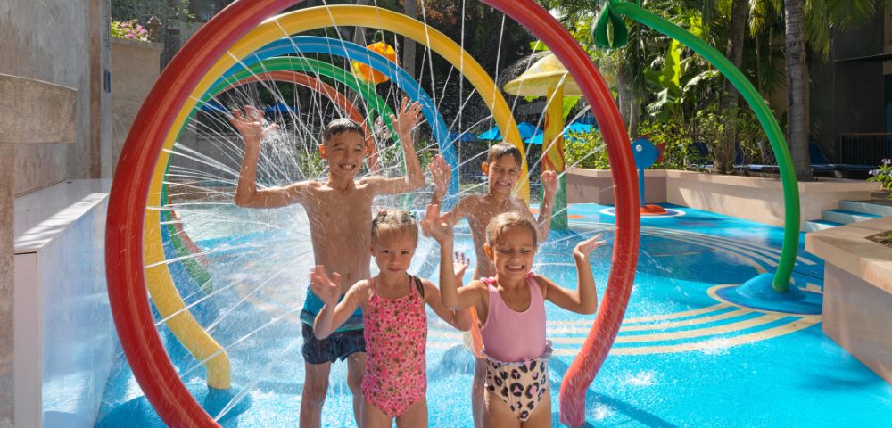 Children joyfully play in a water park, standing under colorful, rainbow-shaped sprinklers. They are smiling, with arms raised amidst splashing water.