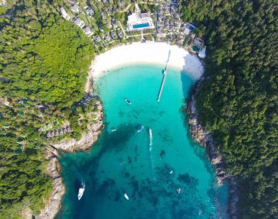 Aerial view of a turquoise bay surrounded by dense green forest. A white sand beach curves along the shore, with boats dotted across the water.