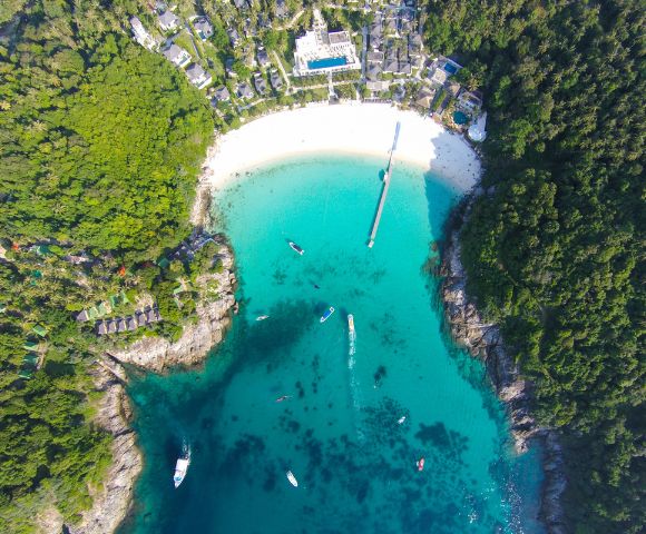 Aerial view of a turquoise bay surrounded by dense green forest. A white sand beach curves along the shore, with boats dotted across the water.