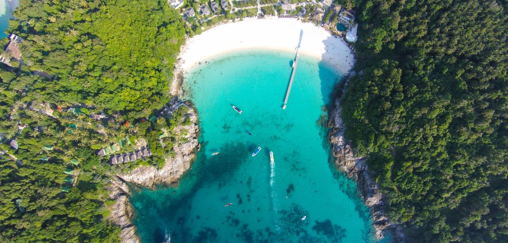 Aerial view of a turquoise bay surrounded by dense green forest. A white sand beach curves along the shore, with boats dotted across the water.