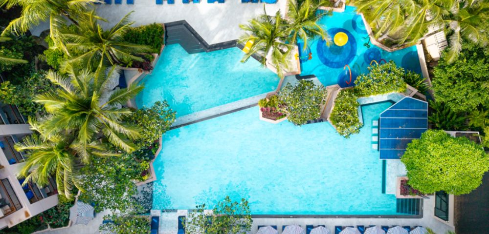 Aerial view of a tropical resort pool surrounded by lush palm trees. Guests relax on blue loungers under umbrellas. The scene is serene and inviting.