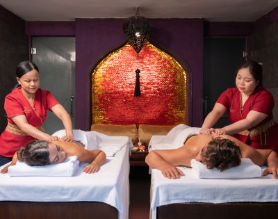 Two people receiving massages on side-by-side tables in a dim, serene spa room with red accents and warm lighting, creating a relaxed atmosphere.