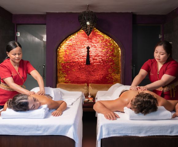 Two people receiving massages on side-by-side tables in a dim, serene spa room with red accents and warm lighting, creating a relaxed atmosphere.