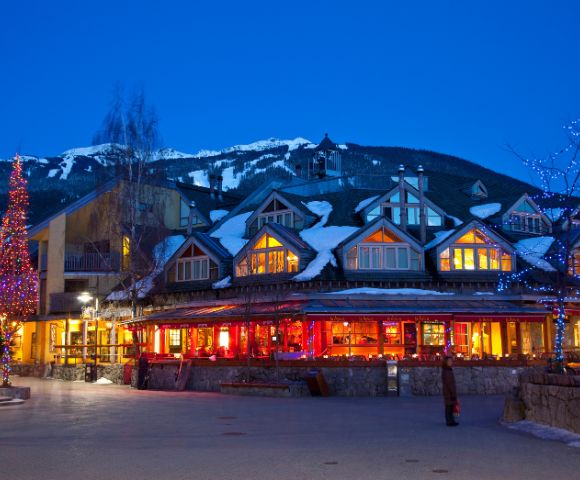 Alpine lodge with warm lights glows at twilight, set against snow-covered mountains. Festive tree and blue-lit trees add a cozy winter charm.
