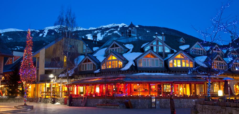 Alpine lodge with warm lights glows at twilight, set against snow-covered mountains. Festive tree and blue-lit trees add a cozy winter charm.
