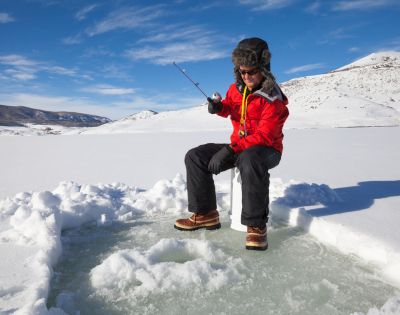A person in a red coat and fur hat ice fishing on a frozen lake under a clear blue sky. Snowy hills in the background create a serene winter scene.