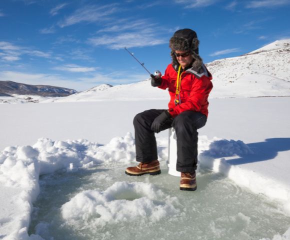 A person in a red coat and fur hat ice fishing on a frozen lake under a clear blue sky. Snowy hills in the background create a serene winter scene.