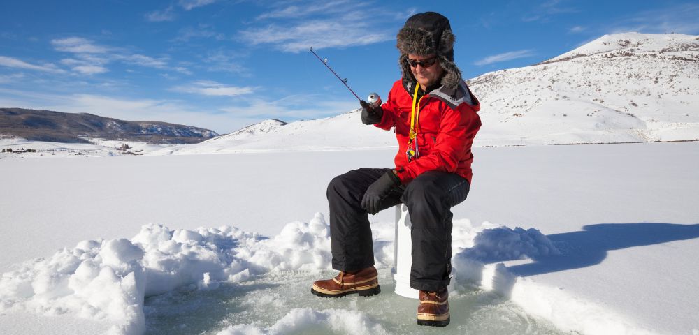 A person in a red coat and fur hat ice fishing on a frozen lake under a clear blue sky. Snowy hills in the background create a serene winter scene.