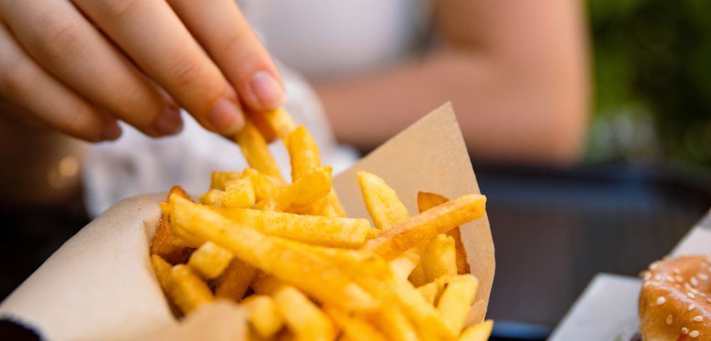 A hand reaches into a basket of golden, crispy French fries on a table, evoking a sense of casual dining. The background is softly blurred.