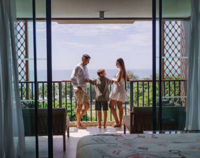 A family of three stands happily on a balcony overlooking a scenic ocean view, with greenery below, under a clear blue sky.