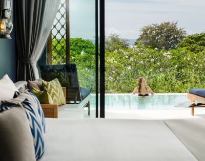 A serene hotel room overlooks a lush garden and pool, where a person relaxes. Bright cushions and an open sliding door create a tranquil, inviting scene.