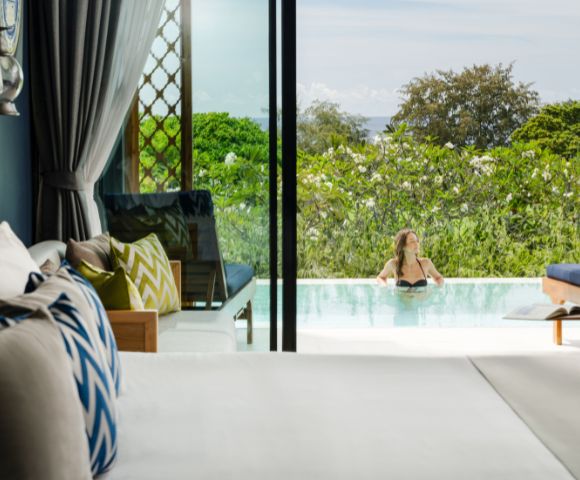 A serene hotel room overlooks a lush garden and pool, where a person relaxes. Bright cushions and an open sliding door create a tranquil, inviting scene.