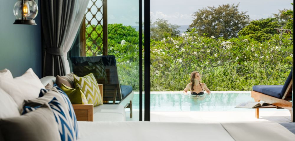 A serene hotel room overlooks a lush garden and pool, where a person relaxes. Bright cushions and an open sliding door create a tranquil, inviting scene.