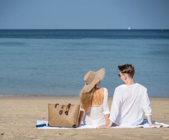 A couple in white outfits sits on a sandy beach, facing the calm sea. A straw hat and handbag are beside them, conveying a peaceful, relaxing vibe.
