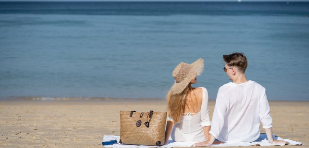 A couple in white outfits sits on a sandy beach, facing the calm sea. A straw hat and handbag are beside them, conveying a peaceful, relaxing vibe.