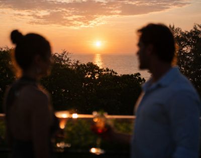 A silhouetted couple enjoys sunset drinks on a balcony overlooking the ocean. Warm hues of orange and pink fill the sky, creating a serene atmosphere.