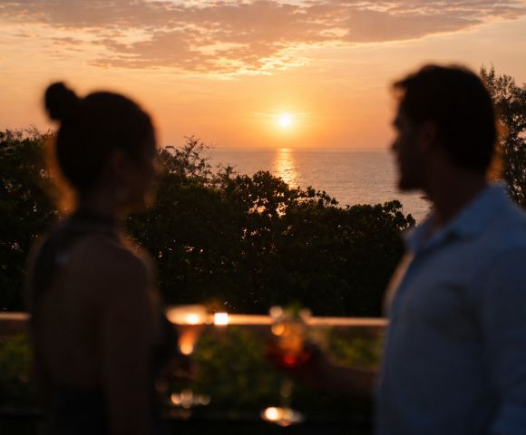 A silhouetted couple enjoys sunset drinks on a balcony overlooking the ocean. Warm hues of orange and pink fill the sky, creating a serene atmosphere.