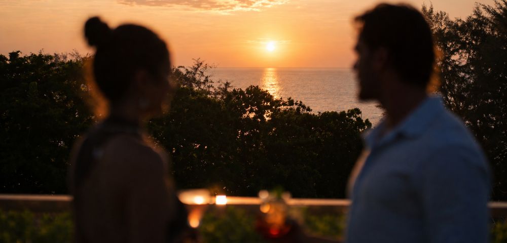 A silhouetted couple enjoys sunset drinks on a balcony overlooking the ocean. Warm hues of orange and pink fill the sky, creating a serene atmosphere.
