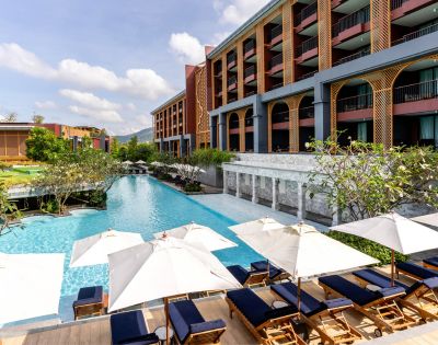 Luxurious hotel pool area with blue water, surrounded by sun loungers under white umbrellas. The modern building has balconies and lush greenery.