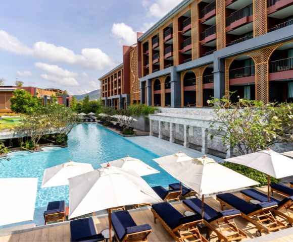 Luxurious hotel pool area with blue water, surrounded by sun loungers under white umbrellas. The modern building has balconies and lush greenery.