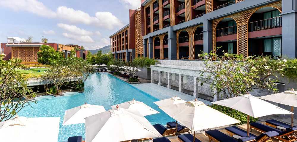 Luxurious hotel pool area with blue water, surrounded by sun loungers under white umbrellas. The modern building has balconies and lush greenery.